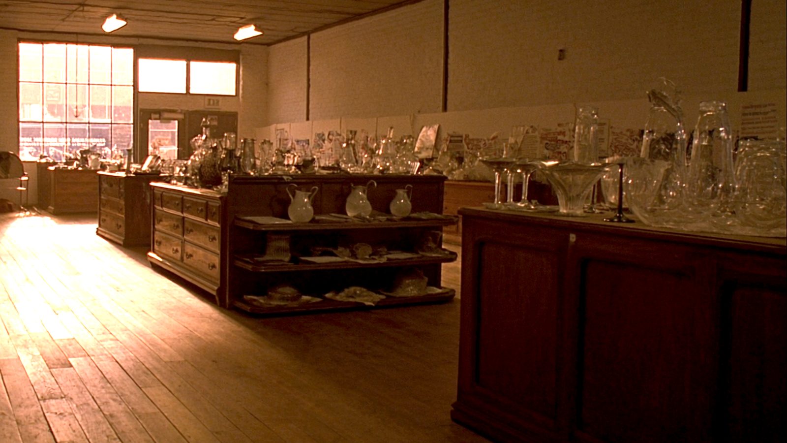 The main aisle on the ground floor late in the afternoon, booths of oak case pieces and pressed glass either side, the overhead lights on and a band of low sun across the floorboards.