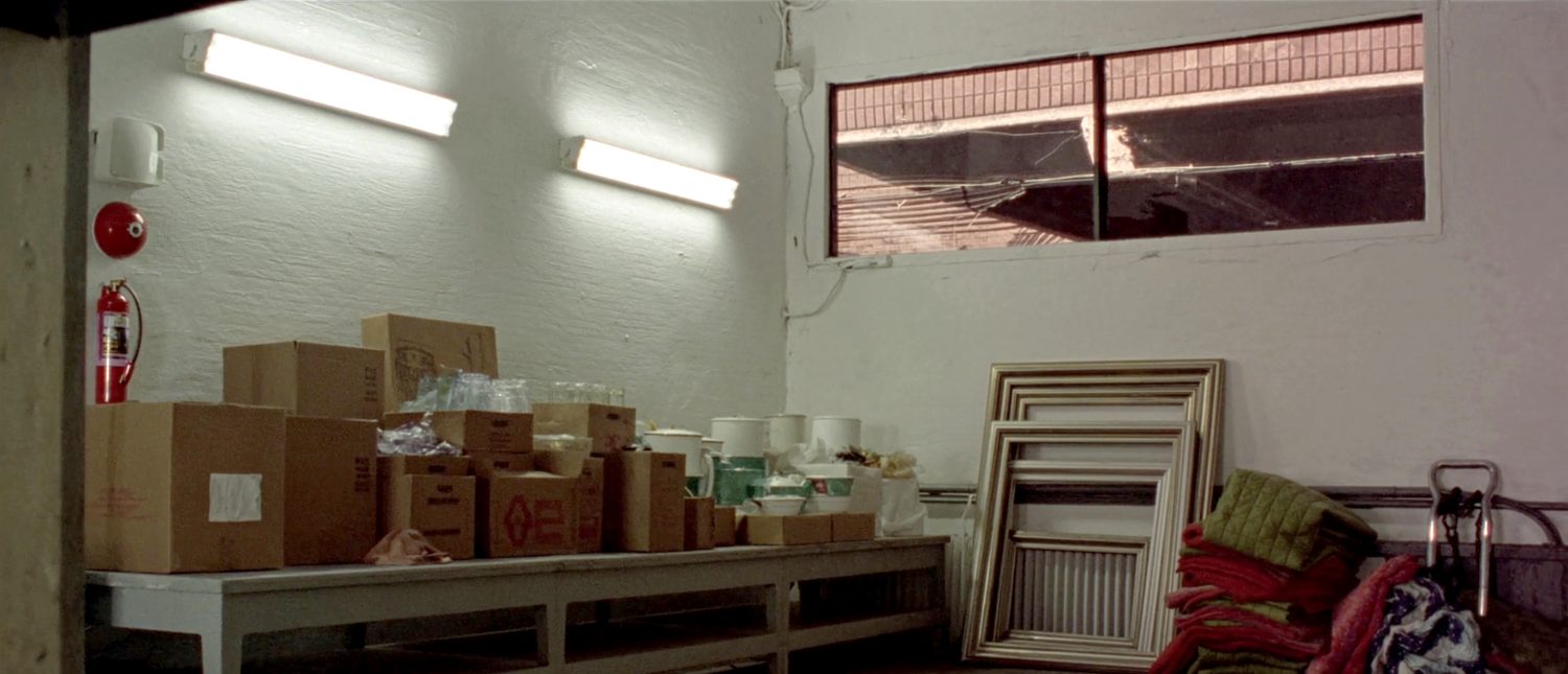 The back sorting room: cartons and crates of glassware and crockery along a long work table, stacked quilts and a row of gilt picture frames leaning against the wall, work lights on overhead.