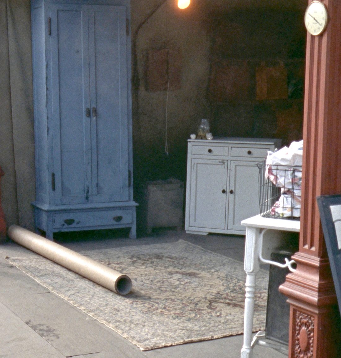 A corner of booth nine: a tall blue painted pine cupboard, a worn rug over the plank floor, a white painted side table with a wire basket of folded linen on it, and a bare bulb lit overhead.