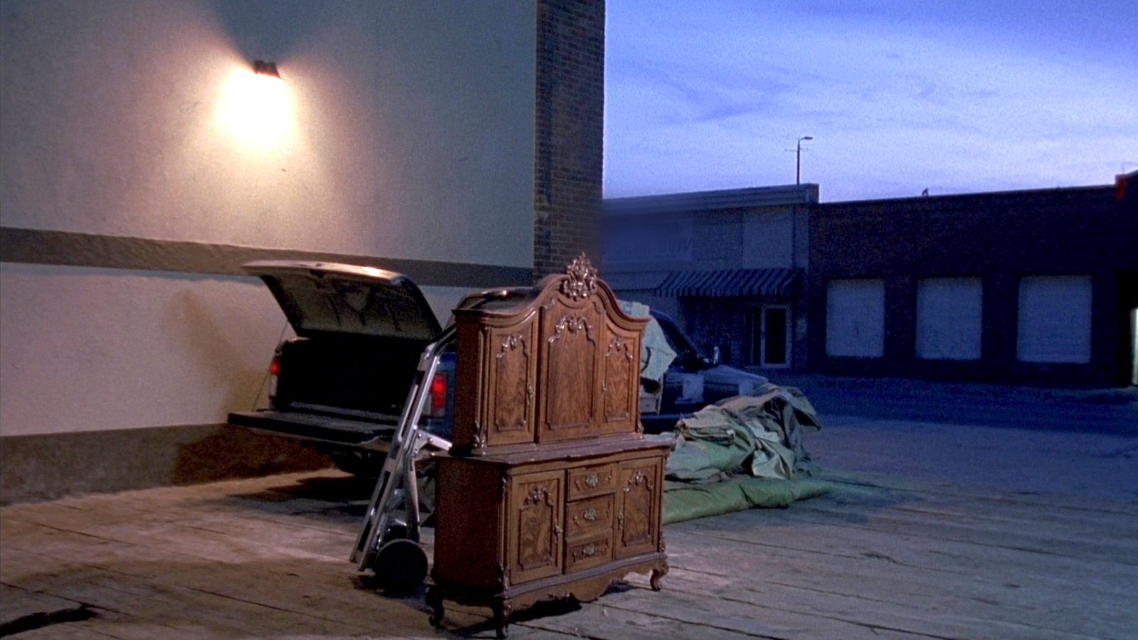 An oak sideboard standing on the wooden loading dock at dusk beside an open pickup tailgate, folded moving blankets and a hand truck alongside, under a lit dock lamp.