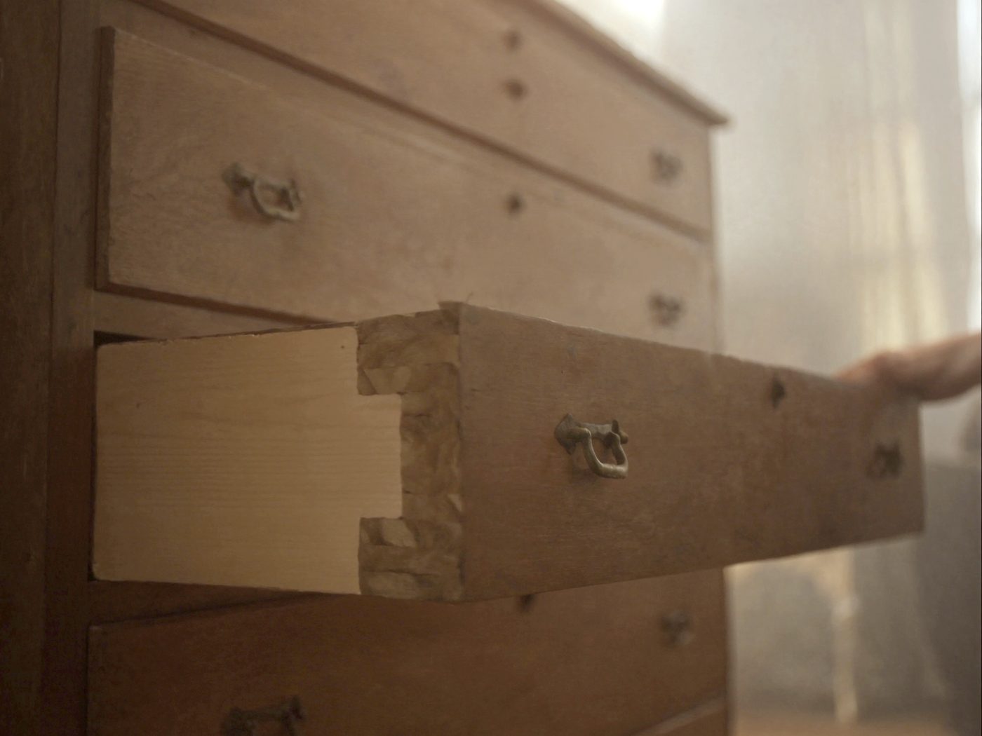 A hand pulling the drawer out of an oak chest far enough to show the hand-cut dovetails at the corner of the bare pine drawer side.