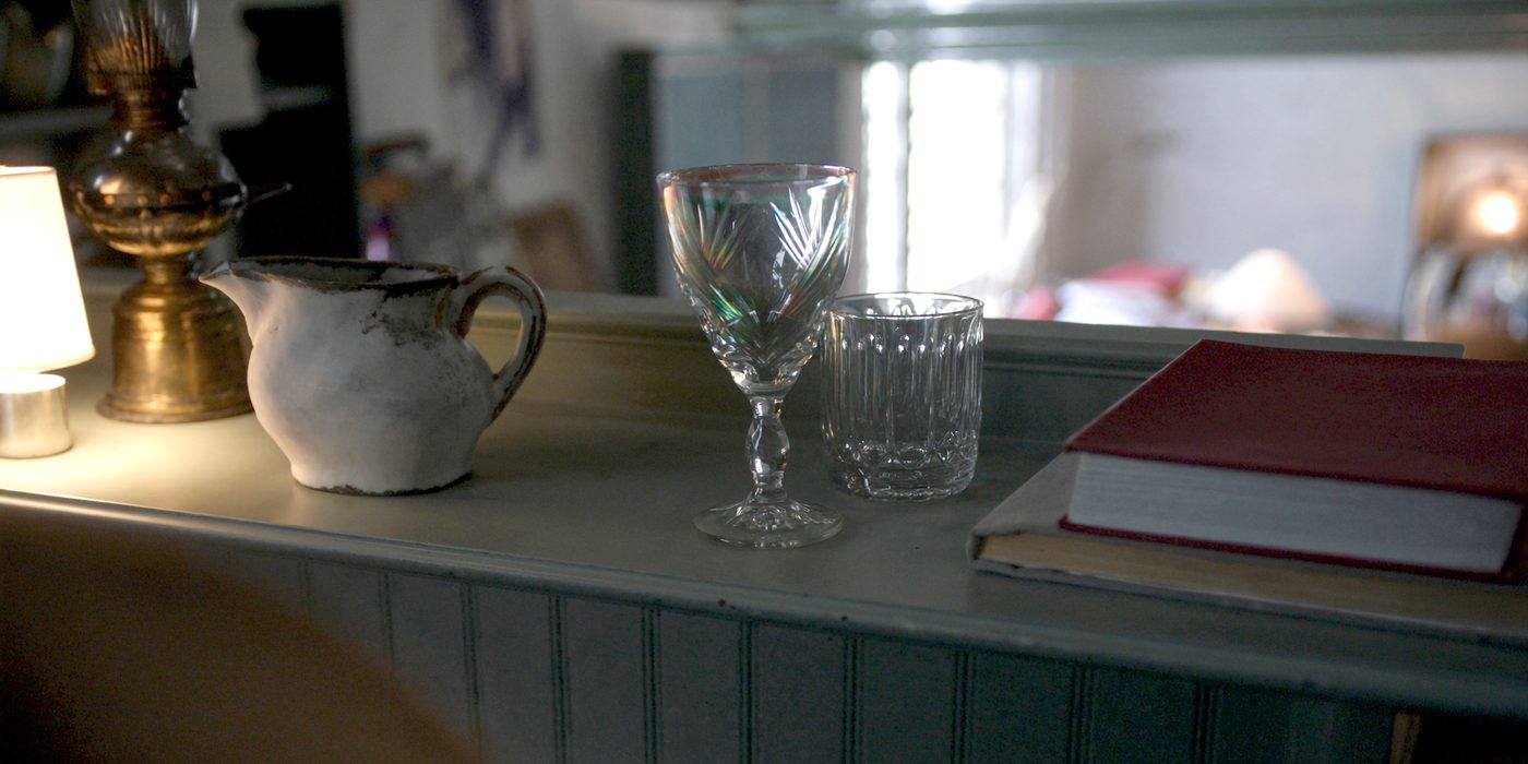 One shelf in booth twenty-two: a cut glass goblet and a ribbed tumbler, an ironstone pitcher, a brass oil lamp and a cloth-bound book, lit by a small lamp just out of frame.